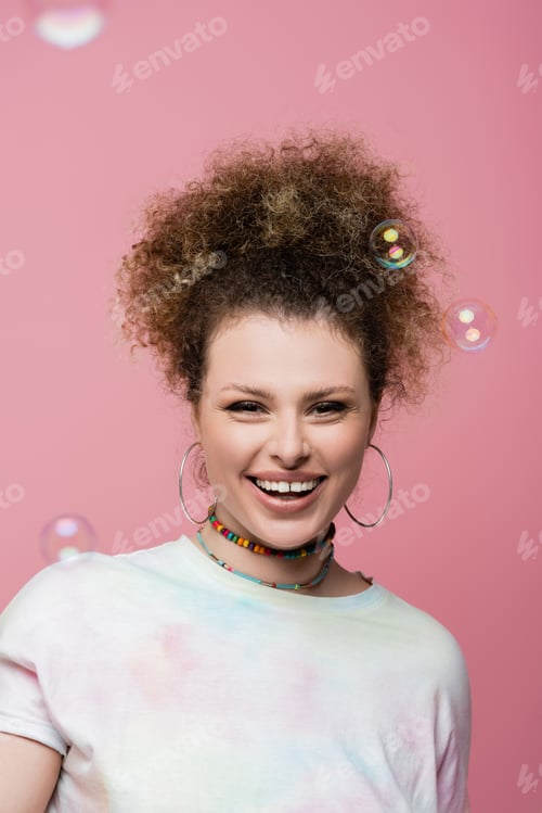 Preview: Positive woman looking at camera near soap bubbles on pink background