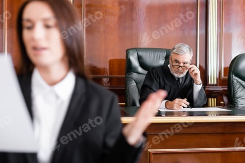 Preview: Grey-haired Judge Adjusting Eyeglasses While Looking at Prosecutor Speaking on Blurred Foreground