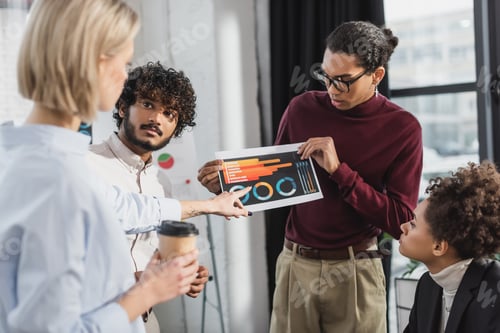 Preview: Blurred businesswoman holding coffee and pointing at paper with graphs near interracial colleagues