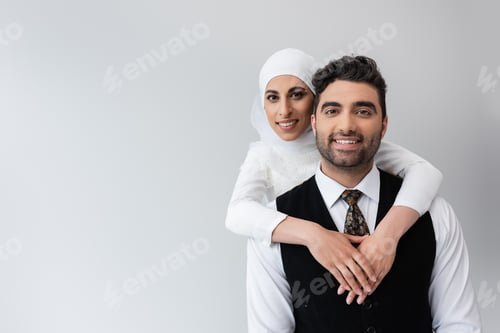 Preview: Happy Muslim Bride in Hijab Hugging Cheerful Groom in Suit Isolated on Grey
