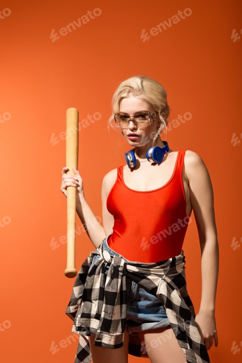 Preview: Confident young woman in vibrant summer outfit holding a baseball bat against a warm backdrop