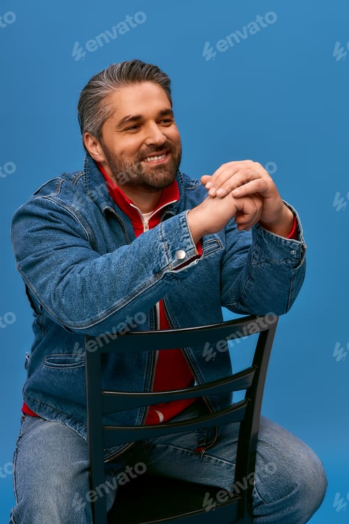 Preview: Stylish man in denim jacket enjoys a relaxed moment against a vibrant blue backdrop