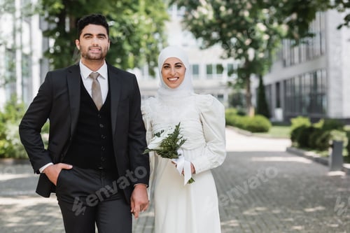 Preview: Portrait of Happy Groom Holding Hands With Smiling Muslim Bride in Hijab With Wedding Bouquet