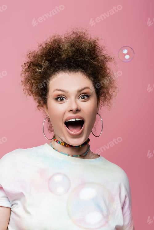 Preview: Excited curly woman looking at camera near soap bubbles on pink background