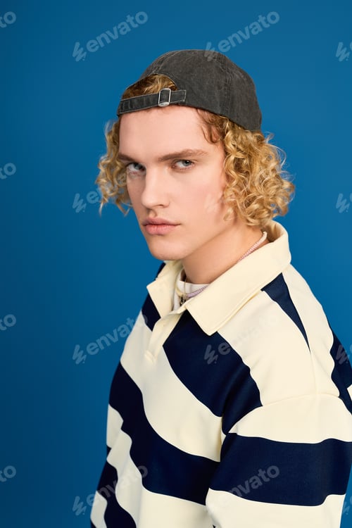 Preview: Curly blonde young man showcasing trendy student style in studio setting
