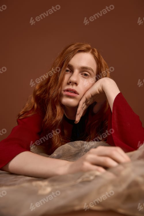 Preview: Handsome young man with long red hair embodies elegance in stylish attire on a warm backdrop