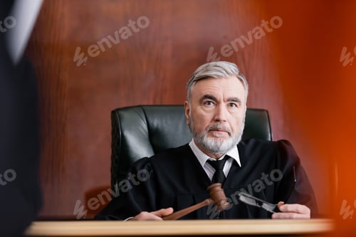Preview: Serious Judge Holding Gavel and Eyeglasses in Court on Blurred Foreground