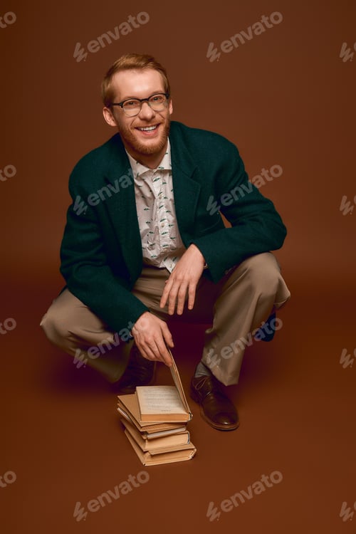 Preview: Stylish red haired man posing with books in a cozy indoor setting