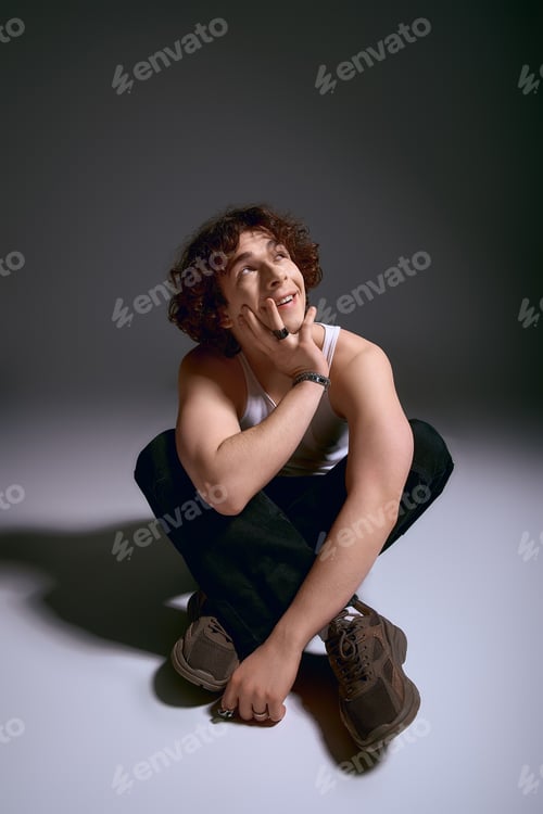 Preview: Young man with curly hair reflects on life while seated in stylish attire against a dark backdrop