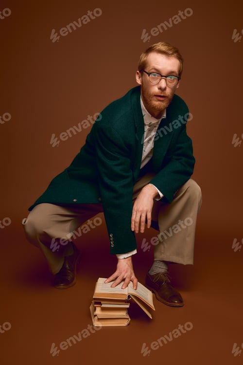 Preview: Handsome red haired man showcasing stylish fashion while balancing on antique books