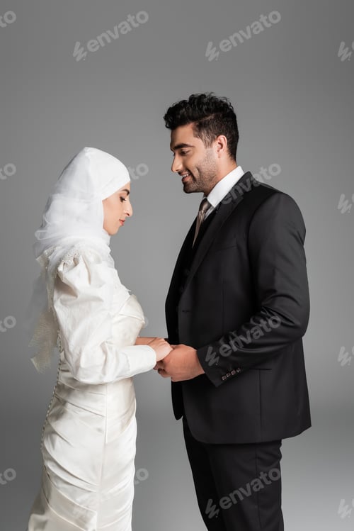 Preview: Muslim Bride in Wedding Dress and Cheerful Groom in Suit Holding Hands Isolated on Grey