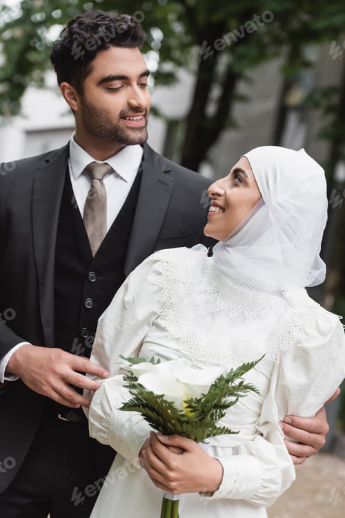 Preview: Cheerful Muslim Bride in Wedding Hijab and White Dress Holding Bouquet and Looking at Groom Outside