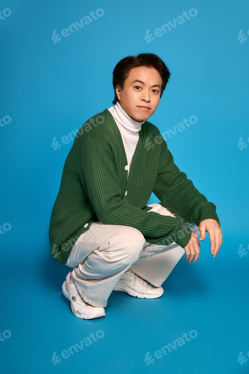 Preview: Stylish young man in green sweater and sneakers expressing unique fashion sense in studio