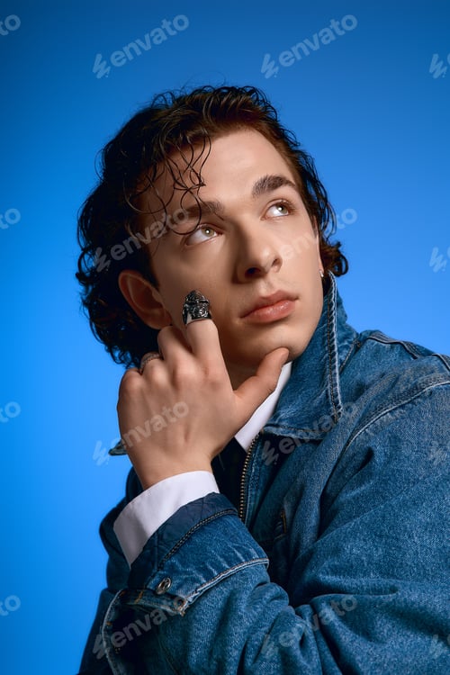 Preview: Young man in denim jacket showcases emotions with curly hair against a vibrant blue backdrop