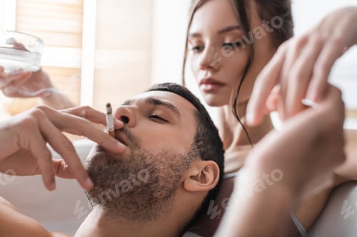 Preview: Bearded man smoking cigarette near blurred woman holding ashtray in bathtub
