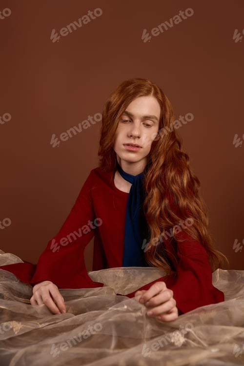 Preview: Young man with long red hair exudes elegance in striking attire against a warm backdrop