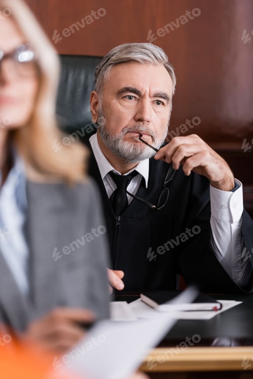 Preview: Thoughtful Judge Holding Eyeglasses Near Advocate on Blurred Foreground