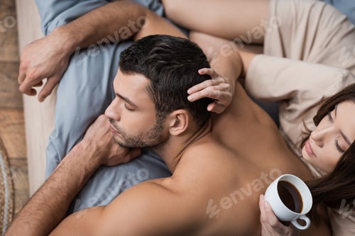 Preview: Overhead view of woman holding coffee and touching hair of muscular boyfriend on bed