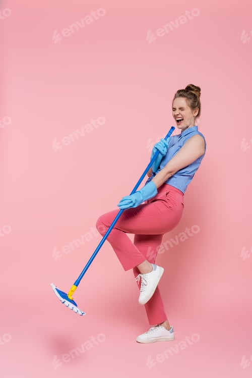 Preview: Full Length of Excited Young Woman in Rubber Gloves Singing While Holding Mop on Pink