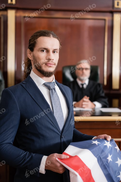 Preview: Attorney Looking at Camera While Holding Usa Flag Near Blurred Judge in Courtroom