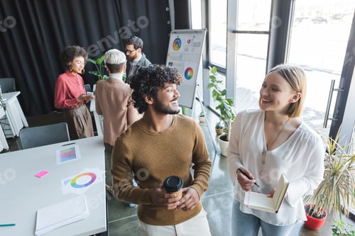 Preview: Cheerful Interracial Advertising Agents With Paper Cup and Notebook Looking at Each Other in Office
