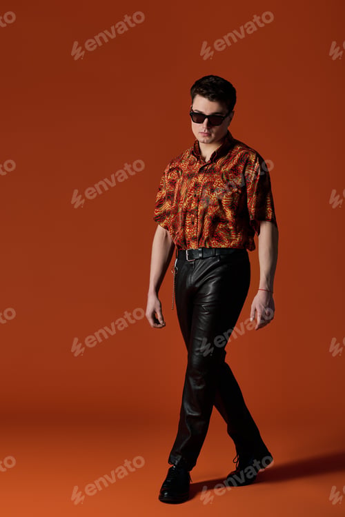 Preview: Stylish young man confidently walks against a vivid orange backdrop showcasing modern fashion