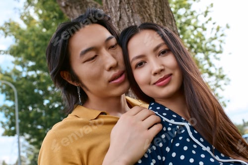 Preview: Young couple sharing an intimate moment in stylish outfits under a lush green tree