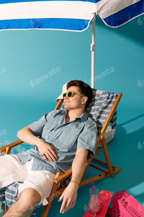 Preview: Stylish young man relaxing under a colorful summer umbrella by the beachside