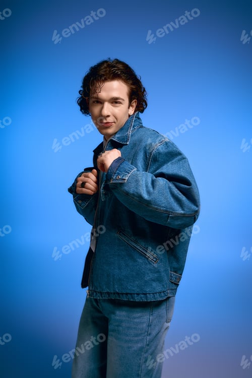 Preview: Emotional young man with curly hair showcases stylish denim against a vibrant blue backdrop