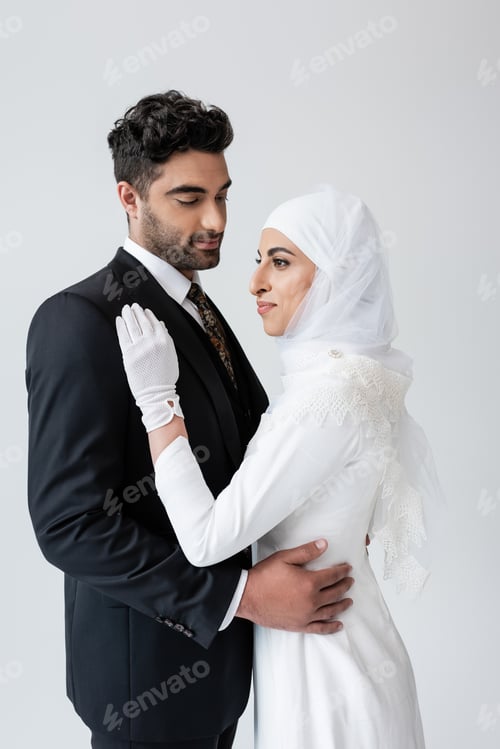 Preview: Young Muslim Bride in Hijab and Wedding Dress and Groom Hugging Isolated on Grey