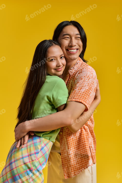 Preview: Young couple embraces joyfully, showcasing their stylish outfits against a vibrant yellow backdrop