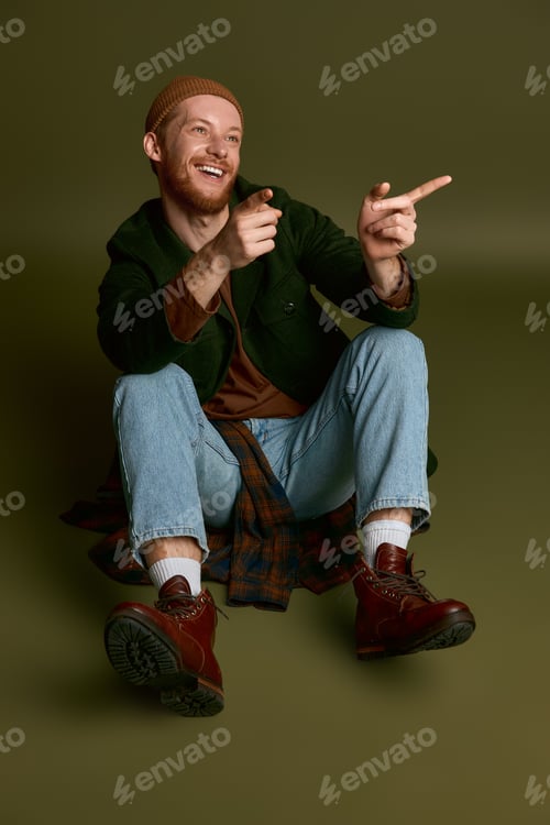 Preview: Stylish young man exudes charm and joy while playfully pointing in a creative indoor setting