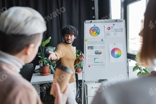 Preview: Curly Indian Man Pointing at Blurred Coworkers Near Flip Chart in Advertising Agency