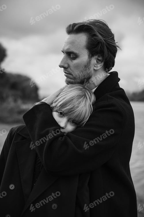 Preview: Father embracing daughter outdoors near water, cloudy sky, monochrome portrait