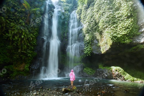 Preview: A young woman in a pink raincoat stands against iconic tropical Fiji Waterfall in Bali