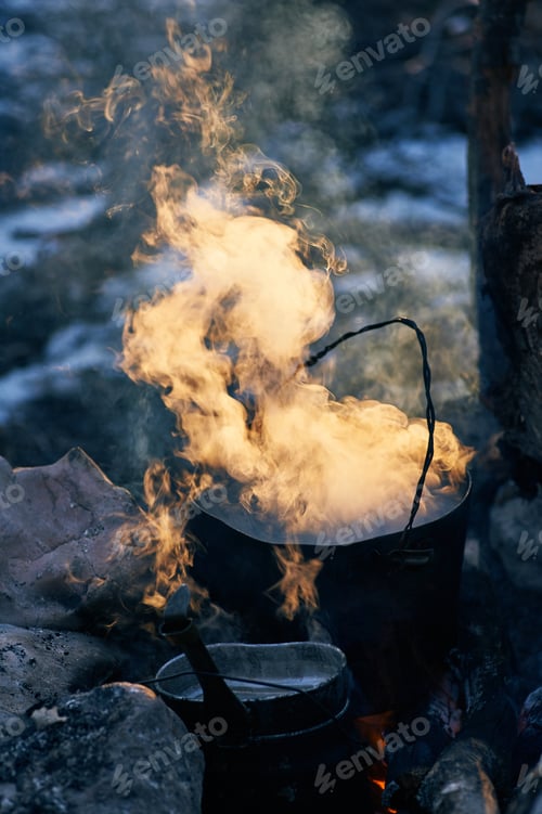 Preview: cooking at campfire. Black charred pot at bonfire with clouds of smoke in sunset lights.