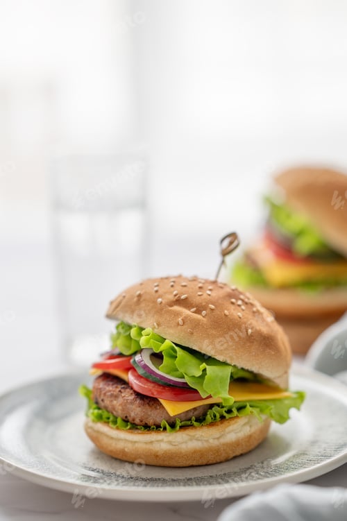 Preview: Two Beef burgers with tomato, cheese, and lettuce on a plate on a table near window with glass of wa