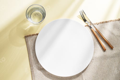 Preview: An empty white plate with glass of water, fork, knife and napkin on a yellow background