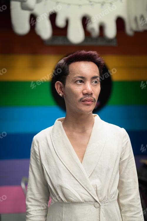 Preview: Non-binary gay Thai man from Isan looking away in front of a rainbow flag wall, capturing authentic