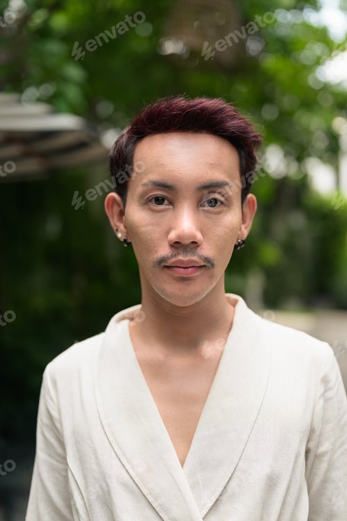 Preview: Vertical portrait of non-binary gay Thai man looking at camera in lush garden courtyard