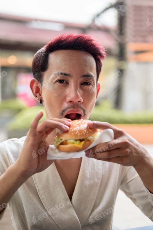 Preview: Thai Non-Binary Gay Man Looking at Camera Eating Burger Outdoors