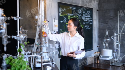 Preview: Young Asian Female Scientist Working with Distillation Equipment in Modern Laboratory with Chalkboar