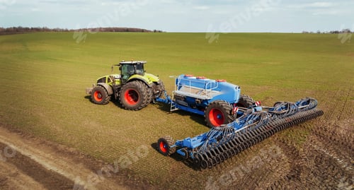 Preview: Agricultural tractor with blue seed drill working on a large green field under a clear sky, showcasi