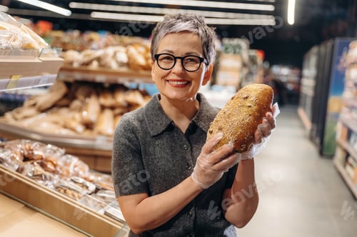 Preview: Mature Asian woman with short gray hair wearing glasses holds a loaf of bread in a bakery aisle, sur
