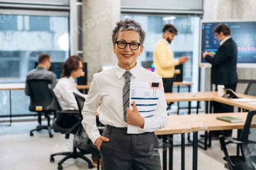 Preview: Confident businesswoman in modern office environment holding documents, smiling at camera, with coll
