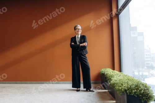 Preview: Businesswoman in black suit stands confidently with arms crossed in modern office space featuring la