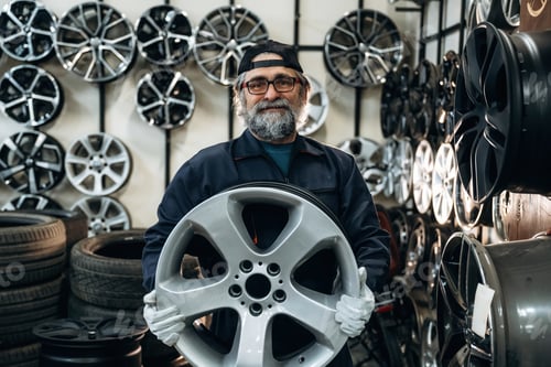 Preview: Older Caucasian male mechanic wearing glasses and gloves holds a large silver alloy wheel in a tire