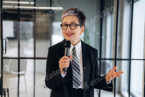 Preview: Businesswoman with short gray hair wearing a black suit and tie speaks confidently into a microphone