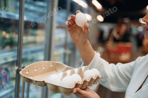 Preview: Woman inspecting fresh eggs in a grocery store, holding one egg above a carton, showcasing quality a