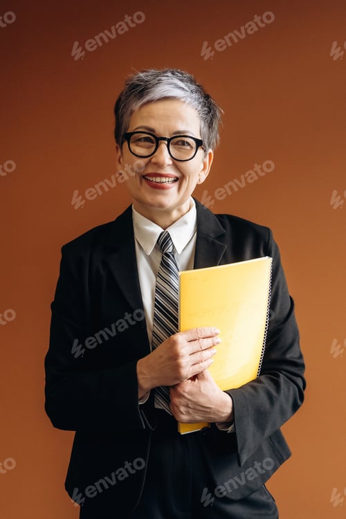 Preview: Businesswoman with short gray hair wearing glasses holds a yellow folder while smiling in a professi